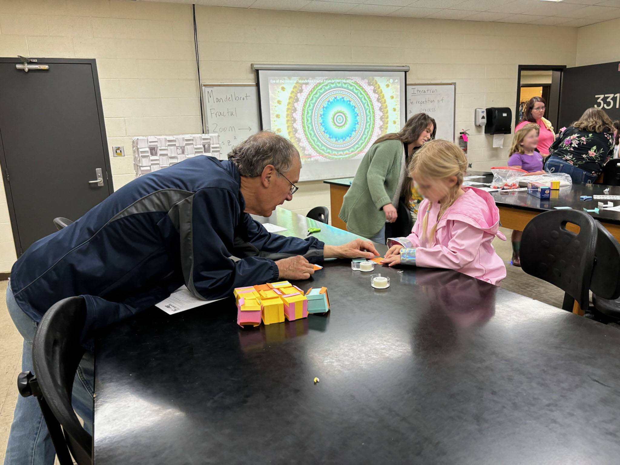 Volunteer reaching across table to help little girl with science project